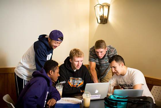 Photo of a group of male Chatham University students huddled around a few laptops and working together