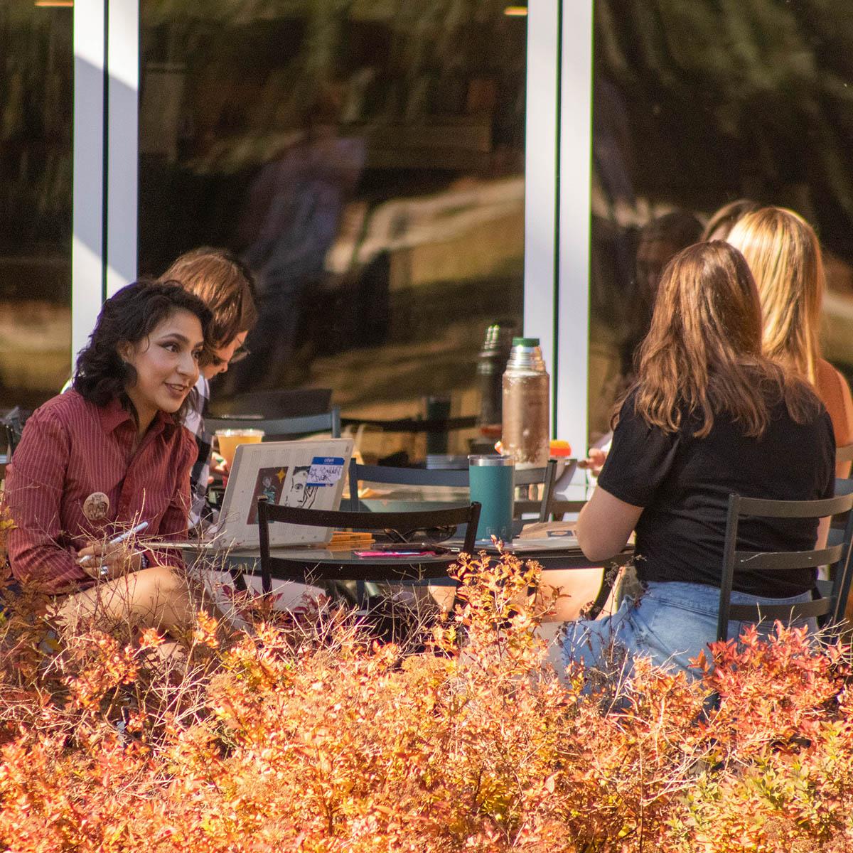Photo of students seated at Cafe Rachel tables outdoors, with autumn foliage in the foreground